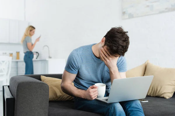 Foyer sélectif de l'homme bouleversé couvrant le visage tout en tenant la tasse près de l'ordinateur portable — Photo de stock