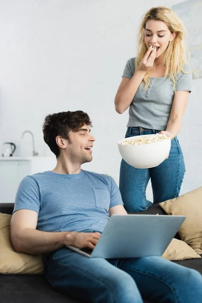 Heureuse fille blonde tenant bol avec pop-corn près de l'homme en utilisant un ordinateur portable — Photo de stock