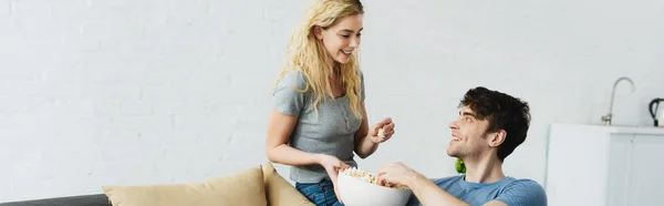 Panoramic shot of happy blonde girl holding bowl with popcorn near man — Stock Photo