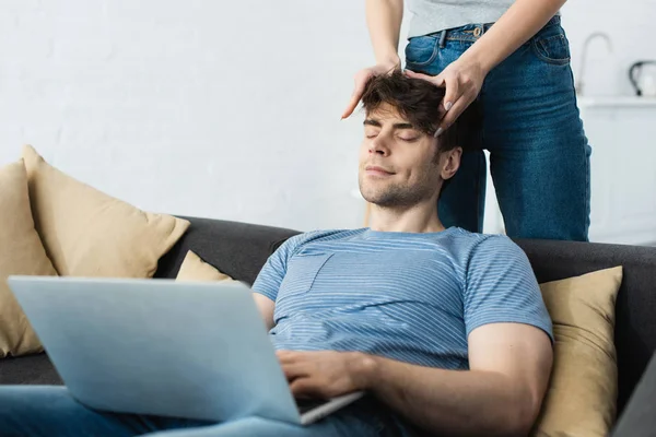 Abgeschnittene Ansicht einer Frau, die den Kopf eines glücklichen Mannes berührt, der mit Laptop auf dem Sofa sitzt — Stockfoto
