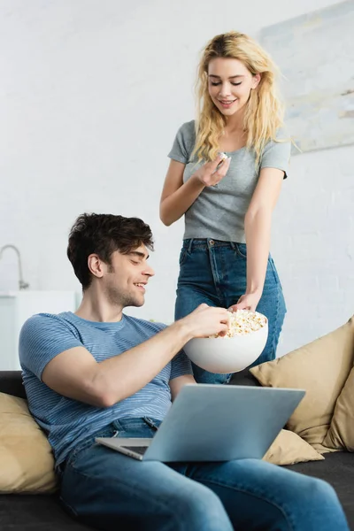 Menina loira feliz segurando tigela com pipoca perto de homem alegre sentado com laptop no sofá — Fotografia de Stock