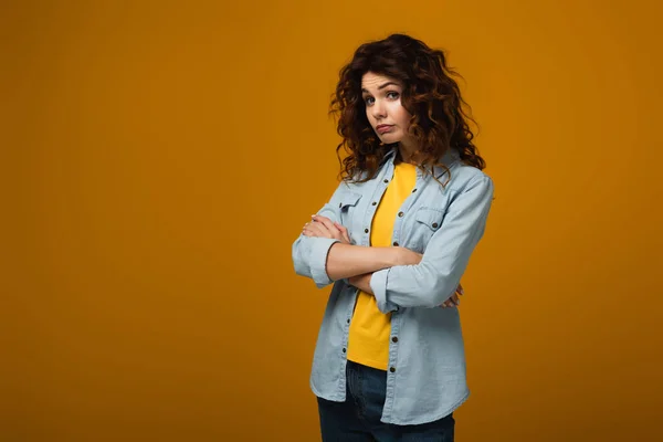 Upset curly redhead woman looking at camera and standing with crossed arms on orange — Stock Photo