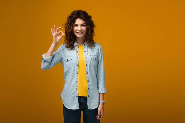 Joyeuse rousse bouclée femme debout et montrant ok signe sur orange — Photo de stock