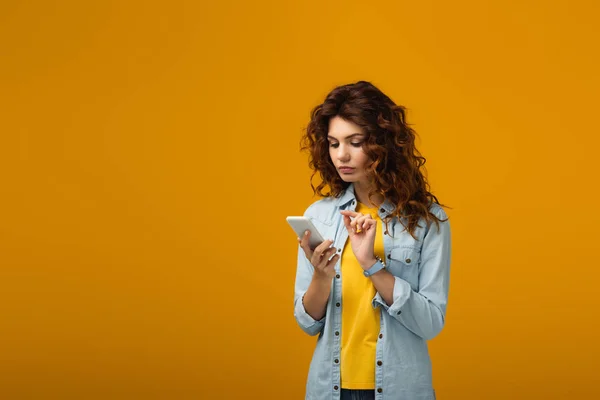 Attractive curly redhead woman looking at smartphone on orange — Stock Photo