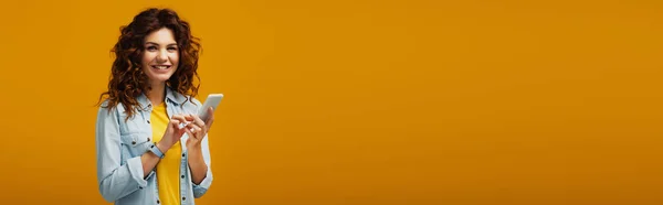 Tiro panorâmico de mulher ruiva encaracolado atraente segurando smartphone em laranja — Fotografia de Stock