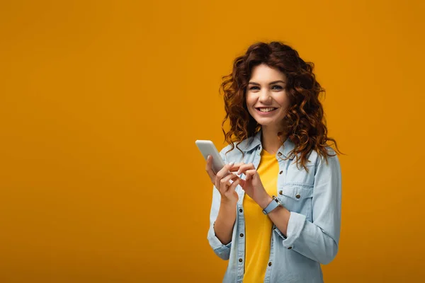 Happy curly redhead woman holding smartphone on orange — Stock Photo
