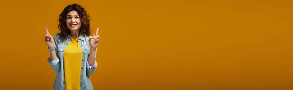 Панорамний знімок щасливої кучерявої рудої жінки, що стоїть з пальцями, схрещеними на апельсині — стокове фото