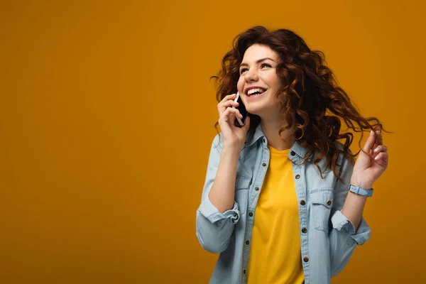 Happy curly redhead woman talking on smartphone and touching hair on orange — Stock Photo