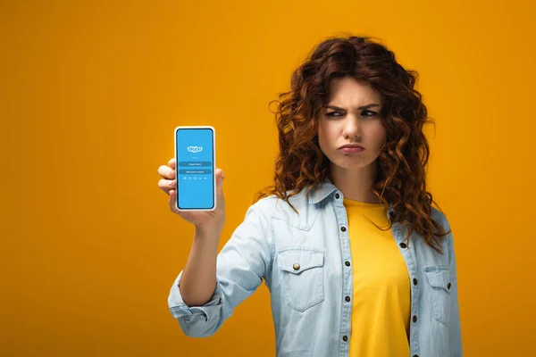 Mujer pelirroja molesta sosteniendo teléfono inteligente con aplicación skype en la pantalla en naranja - foto de stock