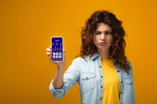 Upset redhead woman holding smartphone with e-health app on screen on orange — Stock Photo