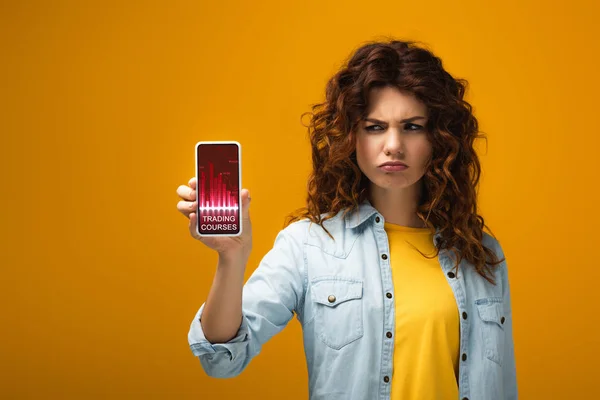 Upset redhead woman holding smartphone with trading courses on screen on orange — Stock Photo