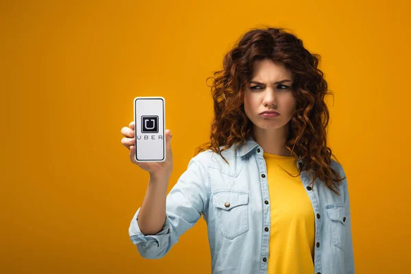 Mujer pelirroja molesta sosteniendo teléfono inteligente con aplicación uber en la pantalla en naranja - foto de stock