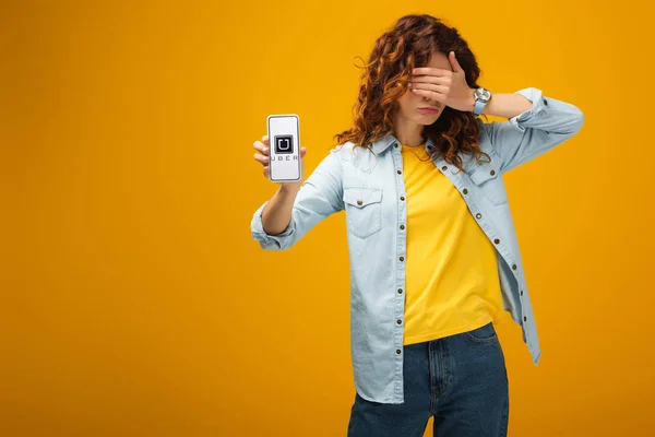 Pelirroja cubriendo los ojos y sosteniendo el teléfono inteligente con la aplicación uber en la pantalla en naranja - foto de stock
