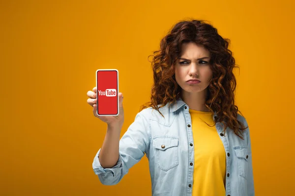 Upset redhead woman holding smartphone with youtube app on screen on orange — Stock Photo