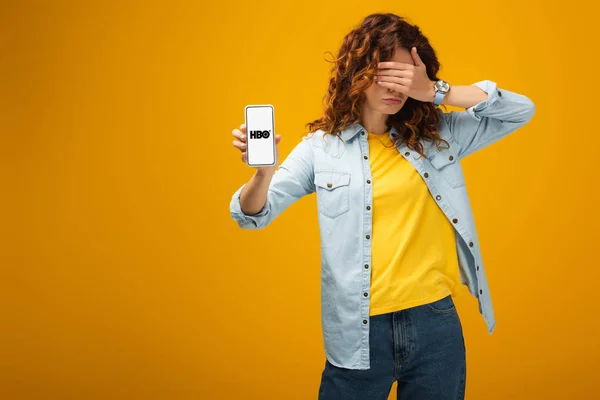 Mulher ruiva cobrindo olhos e segurando smartphone com aplicativo hbo na tela em laranja — Fotografia de Stock