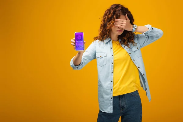 Mujer pelirroja cubriendo los ojos y sosteniendo el teléfono inteligente con e aplicación de compras en pantalla en naranja - foto de stock