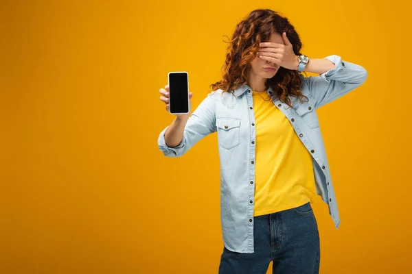 Руда жінка прикриває очі і тримає смартфон з порожнім екраном на помаранчевому — Stock Photo
