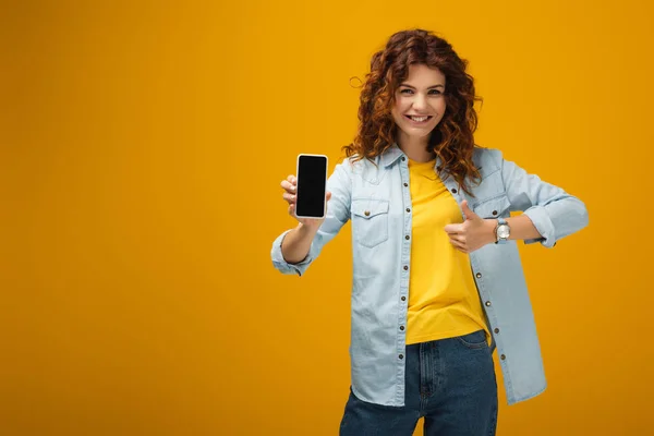 Mujer pelirroja feliz sosteniendo teléfono inteligente con pantalla en blanco y mostrando el pulgar hacia arriba en naranja - foto de stock