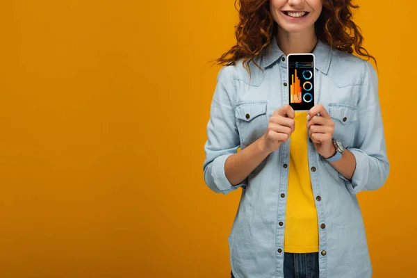 Cropped view of cheerful curly woman holding smartphone with charts and graphs on screen on orange — Stock Photo