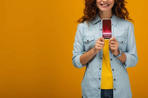 Cropped view of cheerful curly woman holding smartphone with trading courses on screen on orange — Stock Photo