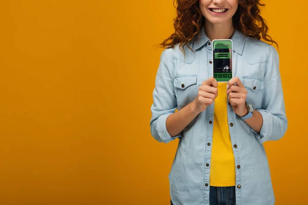 Cropped view of cheerful curly woman holding smartphone with booking app on screen on orange — Stock Photo