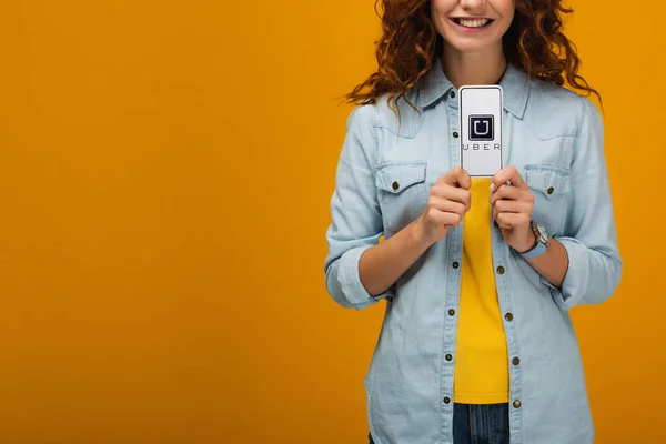 Vista recortada de la mujer rizada alegre sosteniendo teléfono inteligente con aplicación uber en la pantalla en naranja - foto de stock