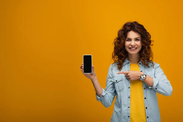 Joyeuse rousse bouclée femme pointant du doigt le smartphone avec écran blanc sur orange — Photo de stock