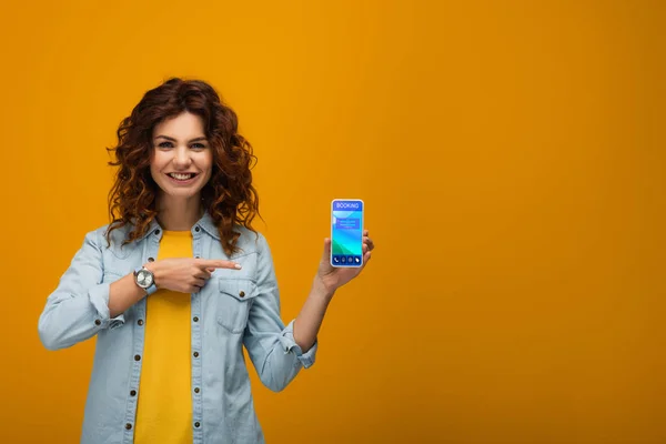 Весела кучерява руда жінка вказує пальцем на смартфон із додатком бронювання на екрані на помаранчевому — стокове фото