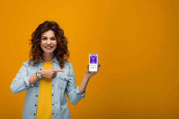 Весёлая кудрявая рыжая женщина, указывающая пальцем на смартфон с приложением itunes на экране на оранжевом — стоковое фото