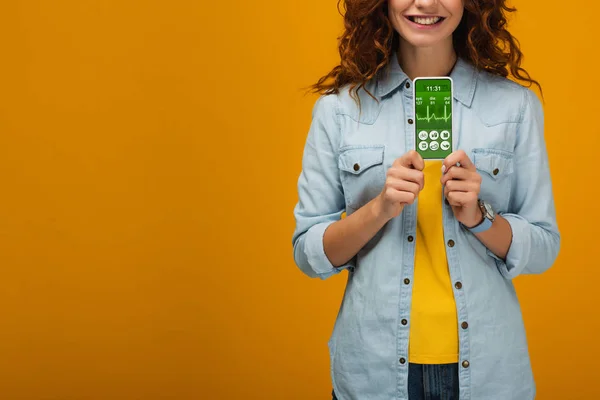Cropped view of cheerful curly woman holding smartphone with e-health app on screen on orange — Stock Photo