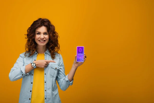 Весела кучерява руда жінка вказує пальцем на смартфон з додатком електронних покупок на екрані на помаранчевому — стокове фото
