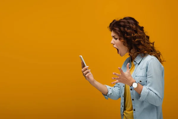 Wütende rothaarige Frau gestikuliert, während sie aufs Smartphone schaut und auf Orange schreit — Stockfoto