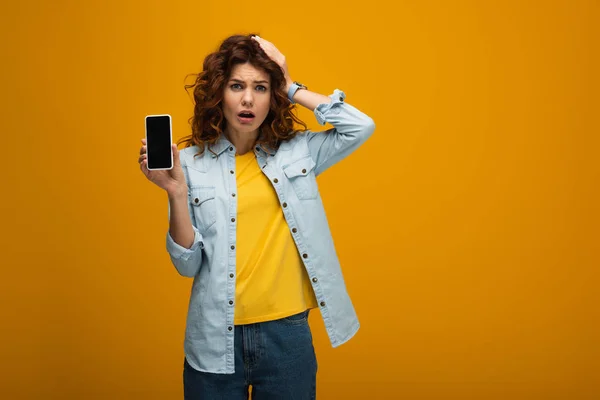 Femme surprise tenant smartphone avec écran vierge et toucher les cheveux sur orange — Photo de stock