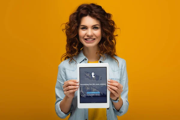 Fröhliche rothaarige Frau hält digitales Tablet mit Tumblr-App auf dem Bildschirm, während sie auf Orange steht — Stockfoto