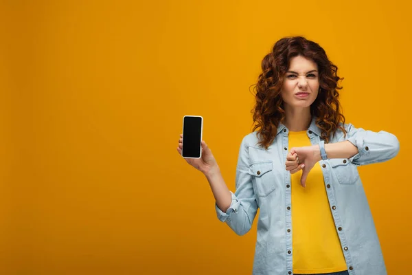 Rousse femme tenant smartphone avec écran blanc et montrant pouce vers le bas sur orange — Photo de stock
