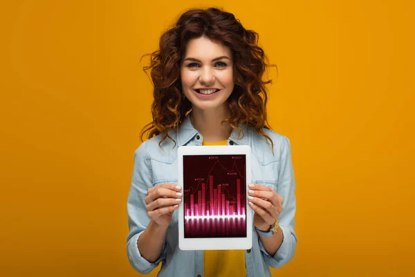 Joyeuse rousse femme tenant tablette numérique avec des graphiques et des graphiques à l'écran tout en se tenant debout sur orange — Stock Photo