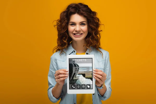 Mujer pelirroja feliz sosteniendo tableta digital con tickets aplicación en la pantalla mientras está de pie en naranja - foto de stock