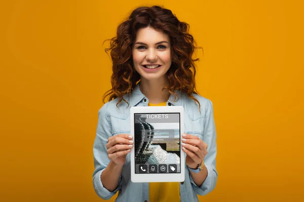 Cheerful redhead woman holding digital tablet with tickets app on screen while standing on orange — Stock Photo