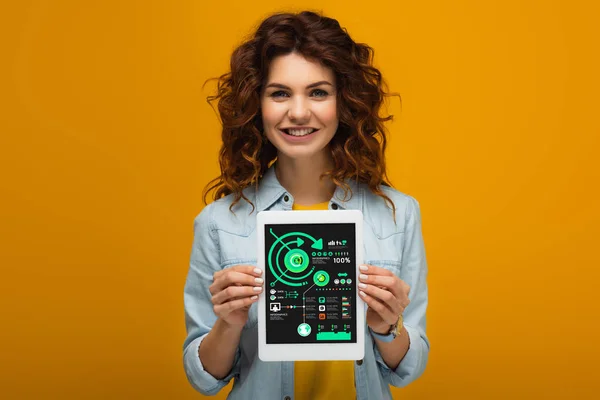 Smiling redhead woman holding digital tablet with charts and graphs on screen while standing on orange — Stock Photo