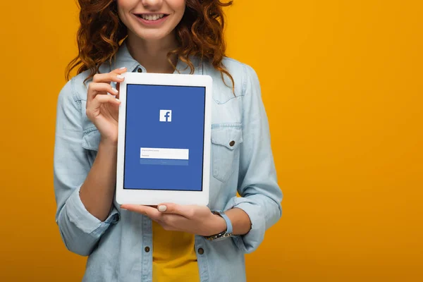 Vista recortada de chica rizada alegre sosteniendo tableta digital con aplicación de facebook en la pantalla aislada en naranja - foto de stock