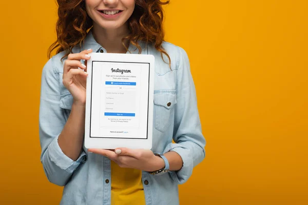 Abgeschnittene Ansicht des fröhlichen lockigen Mädchens mit digitalem Tablet und Instagram-App auf dem Bildschirm isoliert auf orange — Stockfoto