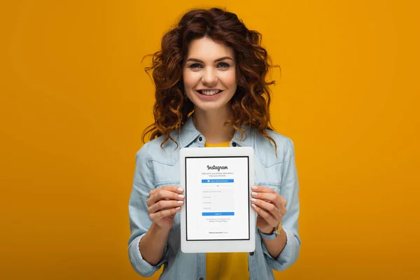 Sonriente pelirroja sosteniendo tableta digital con aplicación instagram en la pantalla mientras está de pie en naranja - foto de stock