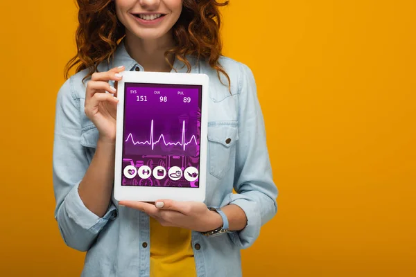 Vista recortada de niña rizada alegre sosteniendo tableta digital con aplicación de salud electrónica en la pantalla aislada en naranja - foto de stock