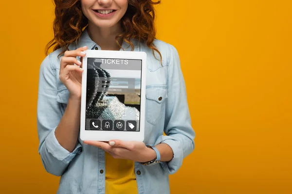 Abgeschnittene Ansicht des fröhlichen lockigen Mädchens mit digitalem Tablet und Ticket-App auf dem Bildschirm isoliert auf orange — Stockfoto