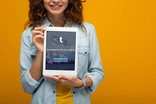 Vista cortada de menina encaracolado alegre segurando tablet digital com aplicativo tumblr na tela isolada na laranja — Fotografia de Stock