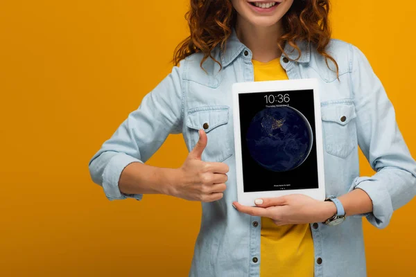 Обрезанный вид веселой кудрявой девушки, держащей цифровой планшет с экраном блокировки и показывающий большой палец, изолированный на оранжевом — стоковое фото
