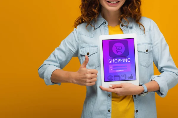 Vista recortada de chica rizada alegre sosteniendo tableta digital con pantalla de aplicación e compras y mostrando el pulgar hacia arriba aislado en naranja - foto de stock