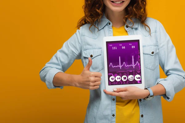 Vista recortada de niña rizada alegre sosteniendo tableta digital con la pantalla de la aplicación de salud e y mostrando el pulgar hacia arriba aislado en naranja - foto de stock