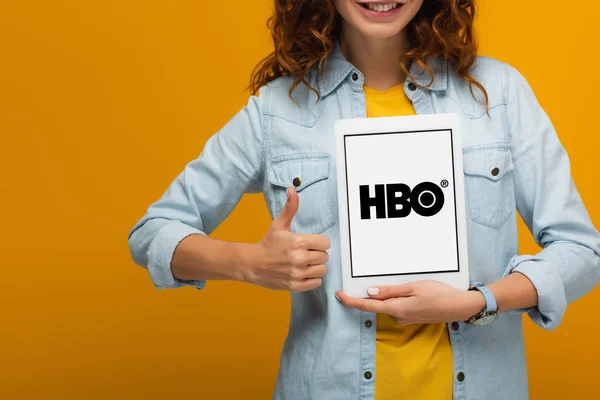 Обрізаний вид Весела Кучерява дівчина тримає цифровий планшет з екрану App HBO і показуючи великий палець до ізольованих на помаранчевий — стокове фото