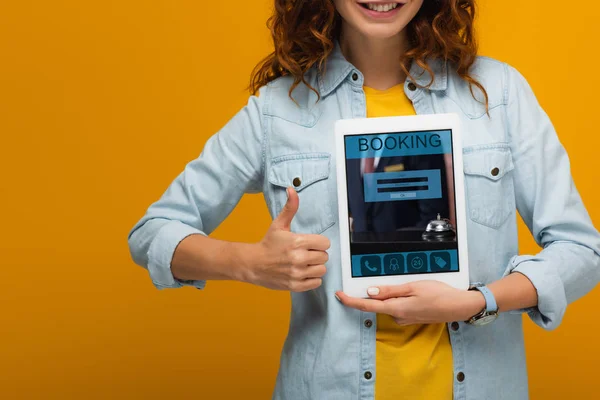 Abgeschnittene Ansicht des fröhlichen lockigen Mädchens, das ein digitales Tablet mit Buchungs-App-Bildschirm hält und den Daumen vereinzelt auf orange zeigt — Stockfoto